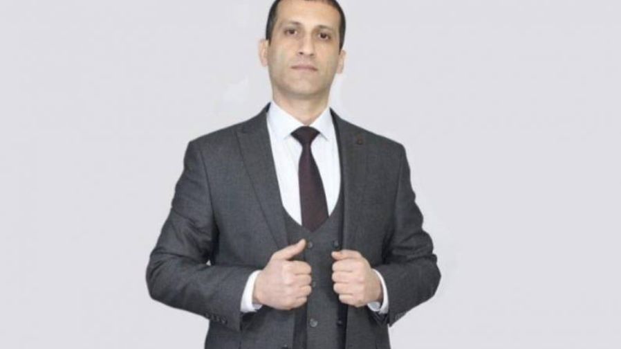 A man in a dark suit, white shirt, and dark tie stands against a plain light background, holding the front of his suit jacket with both hands and looking directly at the camera.