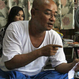 A bald man wearing glasses and a white shirt sits indoors, pointing at a piece of paper. A woman is seated behind him, and patterned curtains are visible in the background.