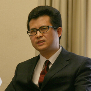 A man wearing glasses and a dark suit with a red tie sits indoors in front of light-colored curtains, looking slightly to the side.