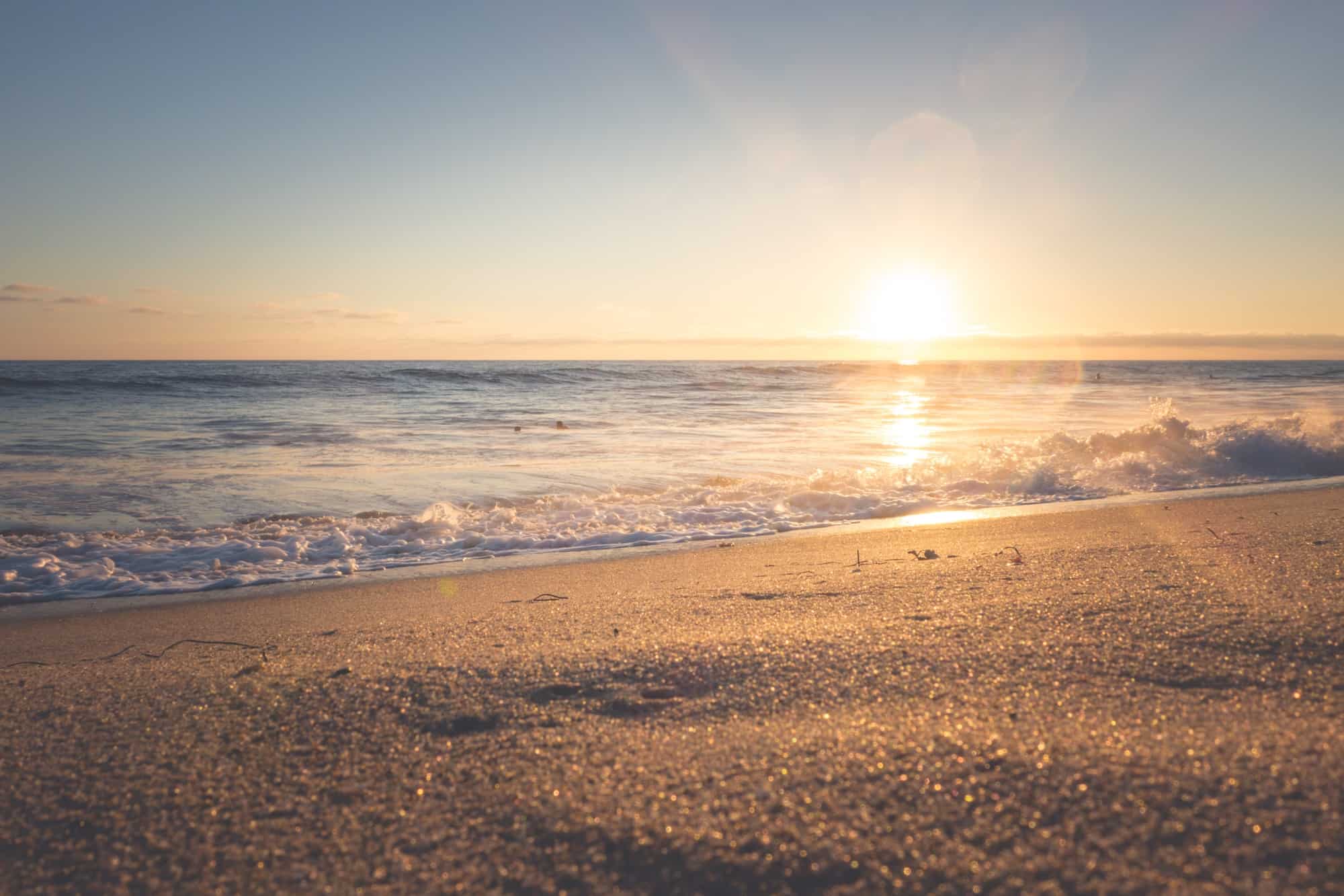 A serene beach scene at sunset, with gentle waves rolling onto the sandy shore and the sun low on the horizon, casting a warm golden glow over the water and sand.