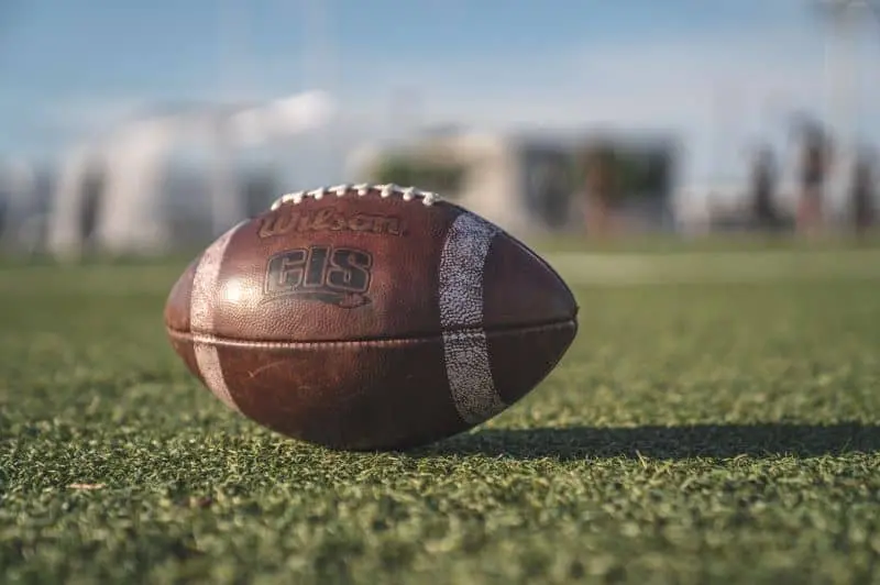 A close-up of an American football resting on a grassy field, with a blurred background of outdoor structures under a clear sky.