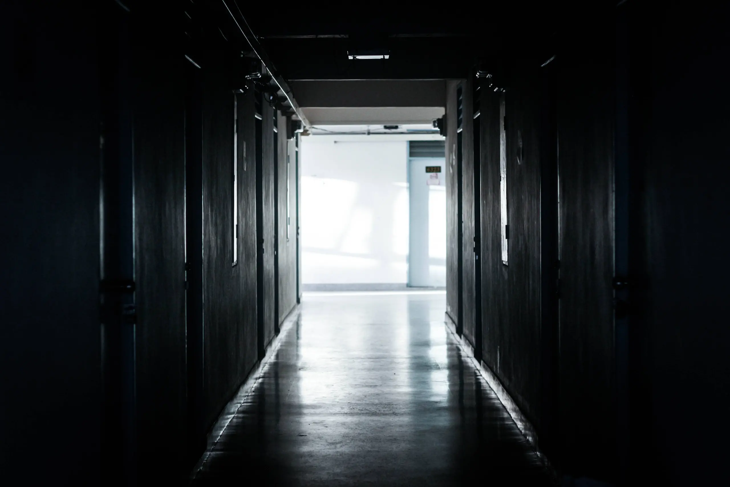 A dark, empty hallway with closed doors on both sides leads to a brightly lit space at the end, where sunlight streams in through windows, illuminating the floor.