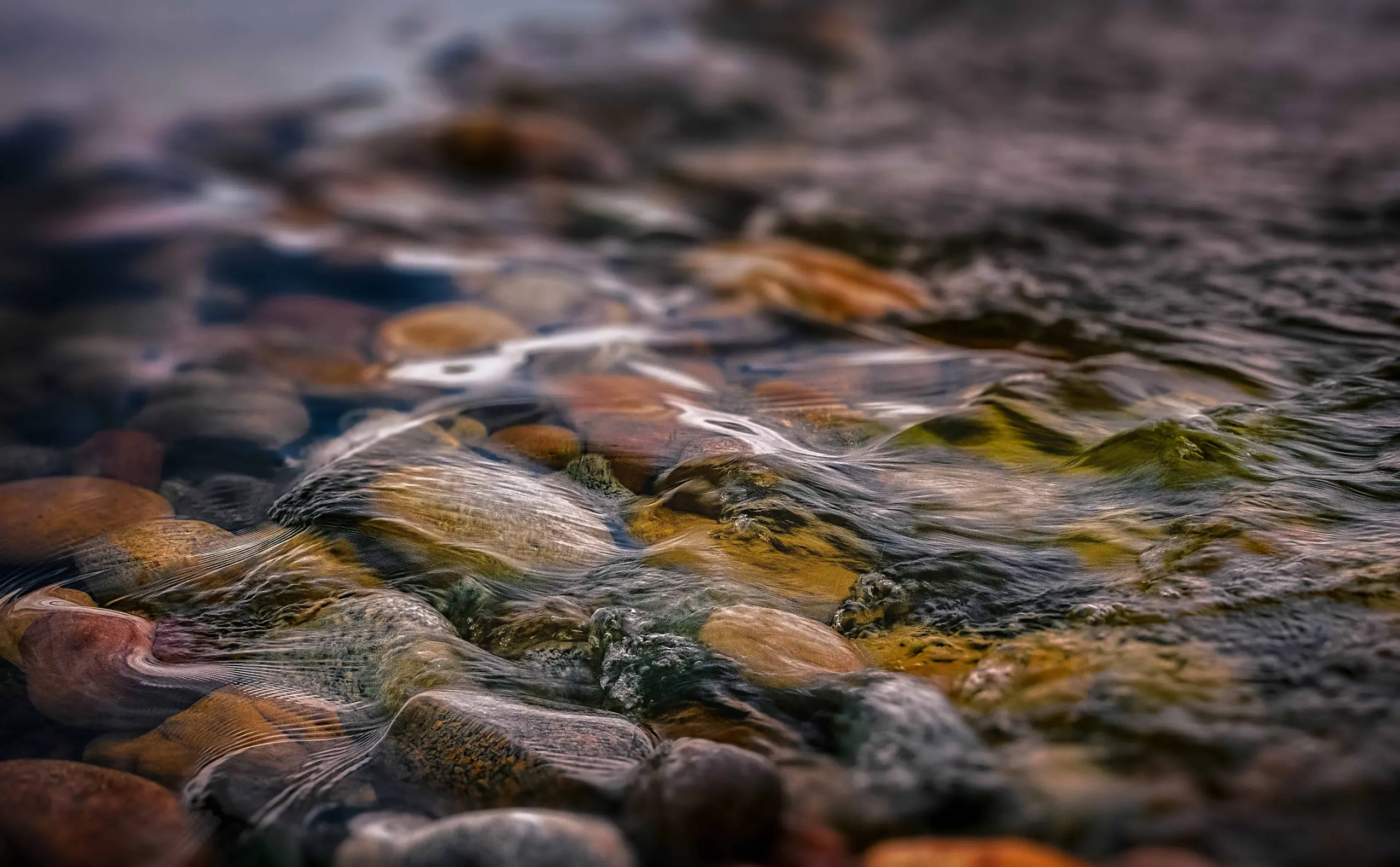 Clear, shallow water flows over smooth, colorful pebbles on a riverbed. The gentle ripples create reflections and distortions, highlighting the earth tones of the stones beneath the surface.