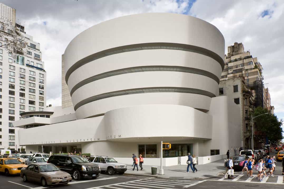 A modern, white, spiral-shaped building stands on a city corner, with taxis and cars on the street and people walking across the crosswalk. Tall buildings surround it under a partly cloudy sky.