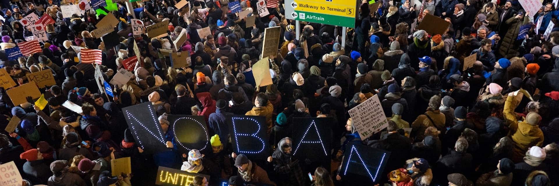 A large crowd of protesters gathers at night, many holding signs including one with illuminated letters spelling “NO BAN.” Some American flags are visible, and people are bundled in coats and hats. A green airport sign is overhead.