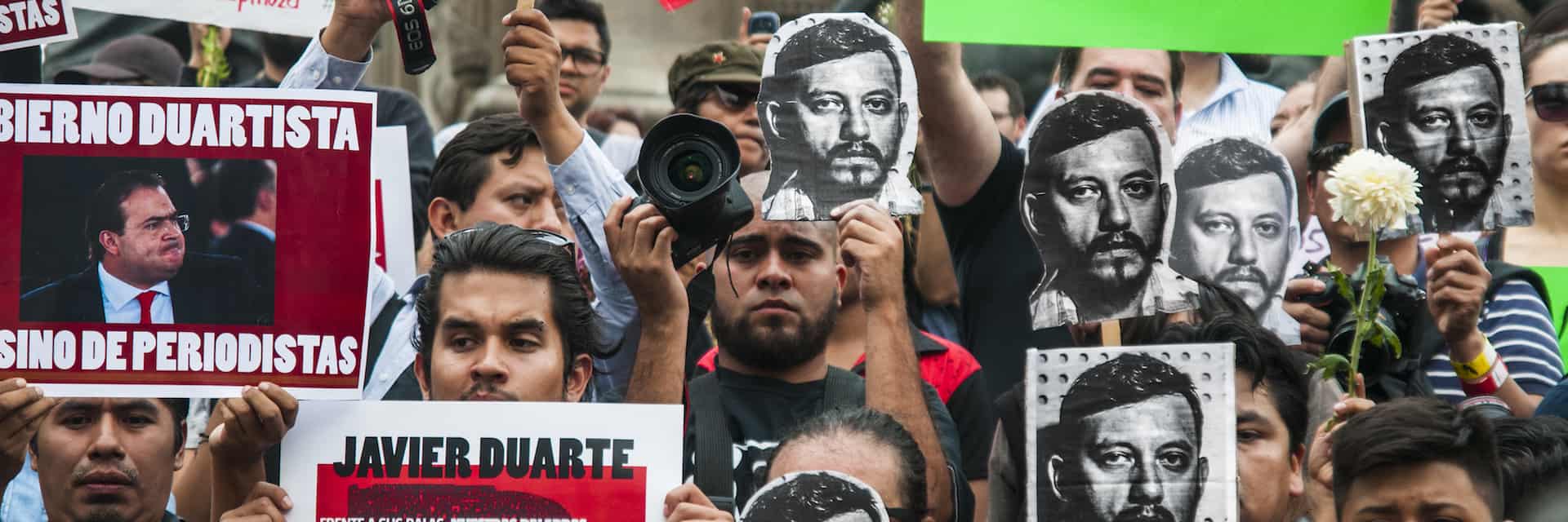 A crowd of protesters holds signs and photos of a man’s face and a placard with another man’s image. Some signs mention “Javier Duarte” and demand justice for journalists. One person holds a white flower.