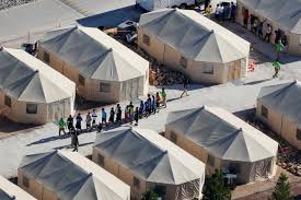 Aerial view of rows of beige tent structures with groups of people standing in lines between them on paved pathways, likely in a temporary camp or shelter setting.