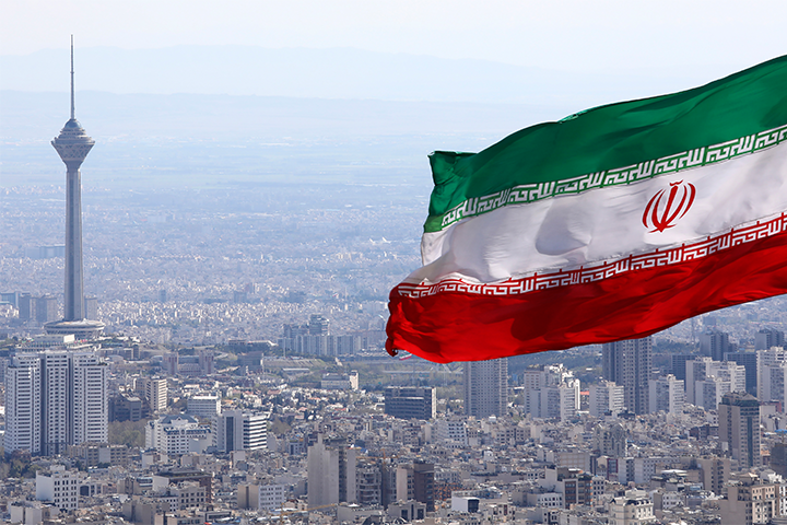 A large Iranian flag waves prominently above the cityscape of Tehran, with the Milad Tower visible in the background among numerous buildings.