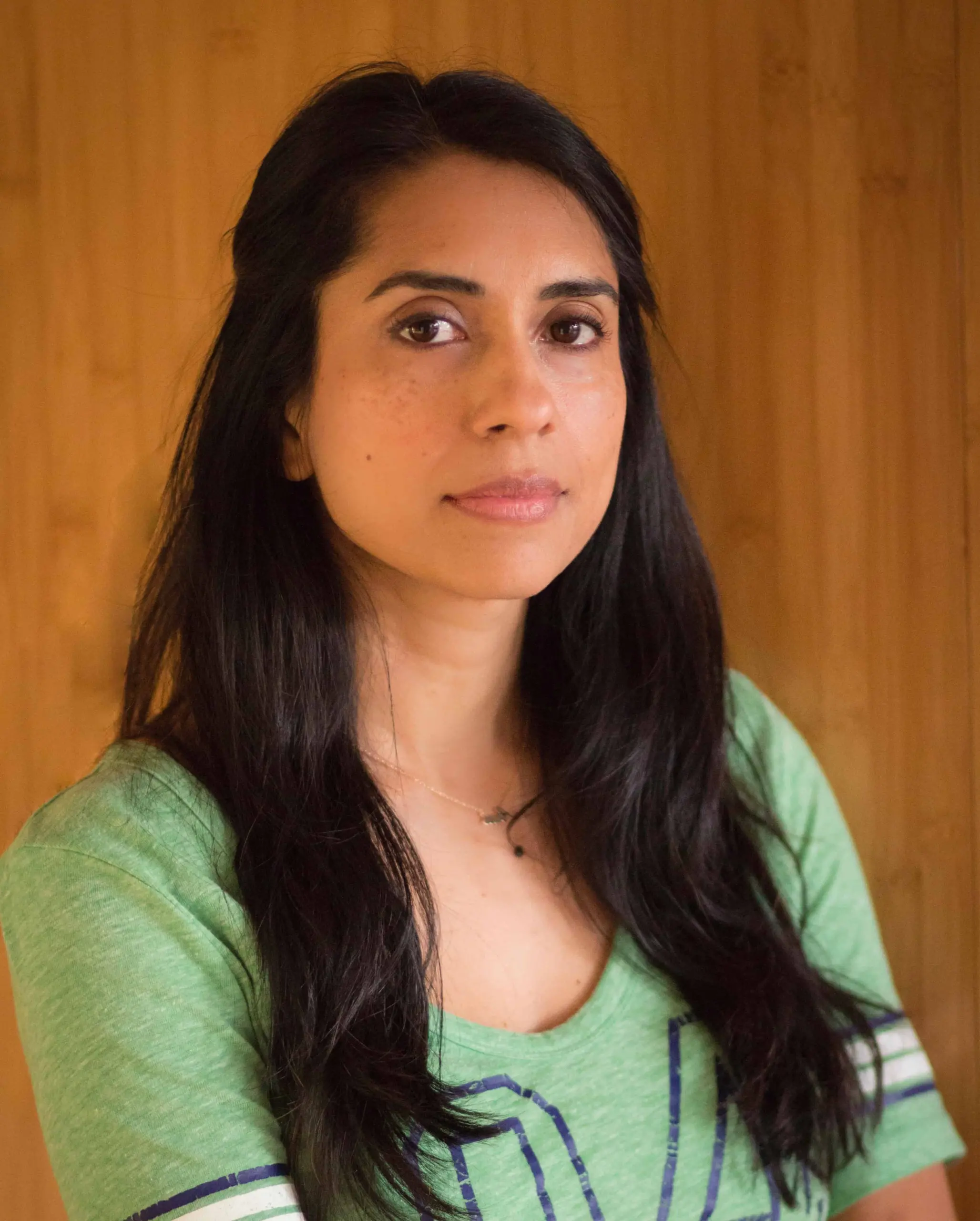 A woman with long dark hair and a neutral expression stands in front of a wooden background, wearing a light green shirt with navy stripes and a delicate necklace.