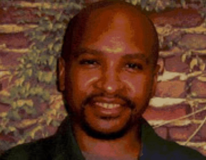 A man with a shaved head and a beard smiles in front of a wall covered in ivy or vines.