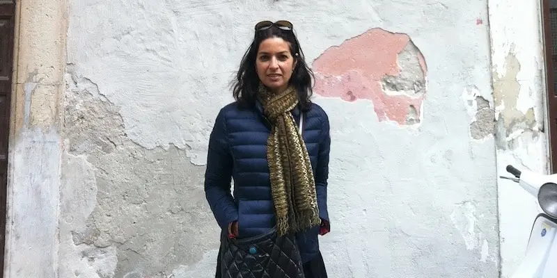 A woman with dark hair, wearing a navy puffer jacket, patterned scarf, and sunglasses on her head, stands in front of a weathered, peeling wall with a white scooter partially visible on the right.