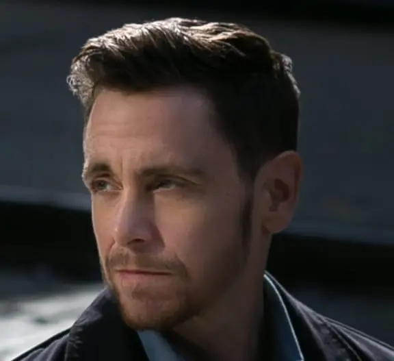 A man with short brown hair and a trimmed beard looks to his left with a serious expression. He is wearing a dark jacket over a light blue shirt, and the background is blurred and dark.