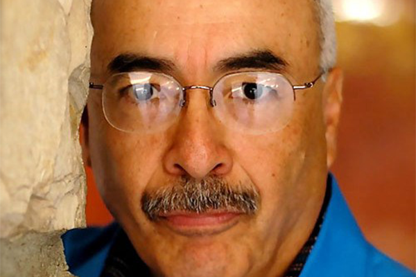 A man with glasses, a mustache, and short gray hair is wearing a blue shirt and looking directly at the camera while standing beside a textured beige wall.