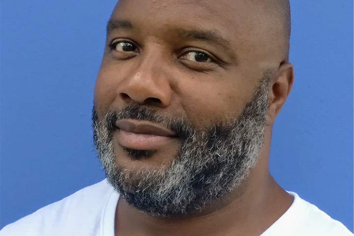 A man with a short, graying beard and shaved head wearing a white shirt stands in front of a solid blue background, looking slightly past the camera with a gentle smile.