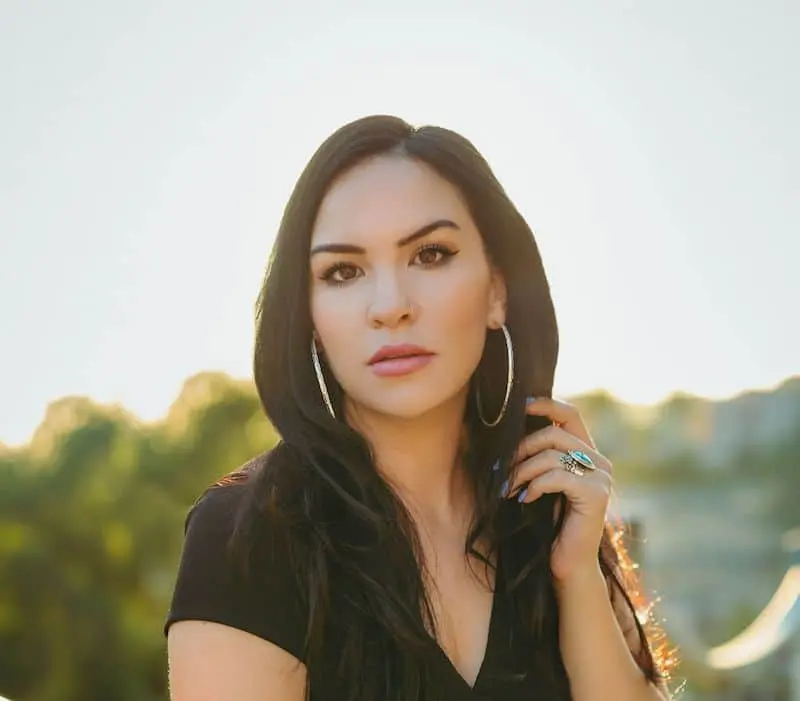 A woman with long dark hair, wearing a black top and large hoop earrings, stands outdoors with sunlight behind her. She looks directly at the camera, with trees and blurred scenery in the background.