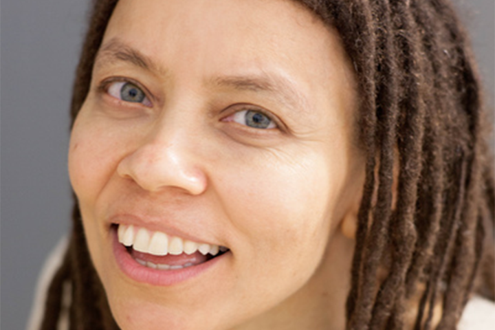 A person with light skin and long brown dreadlocks smiles warmly, showing their teeth. They have light-colored eyes and are looking slightly upward. The background is plain and neutral.