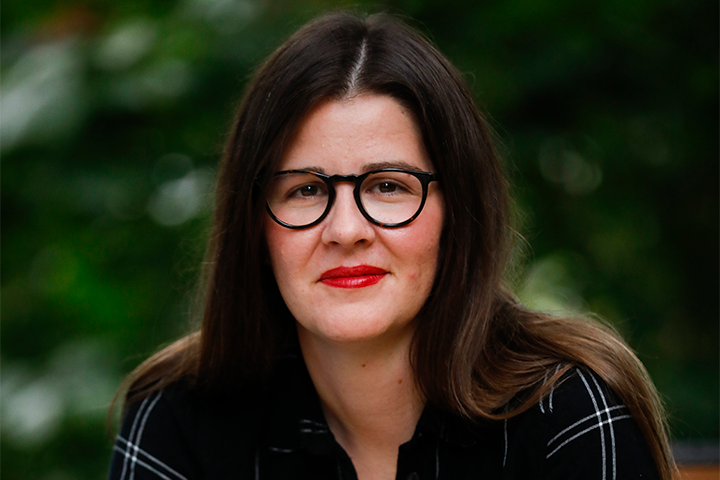 A woman with long brown hair wearing black-rimmed glasses and a black shirt with white lines sits outdoors, smiling slightly. The background is blurred greenery.