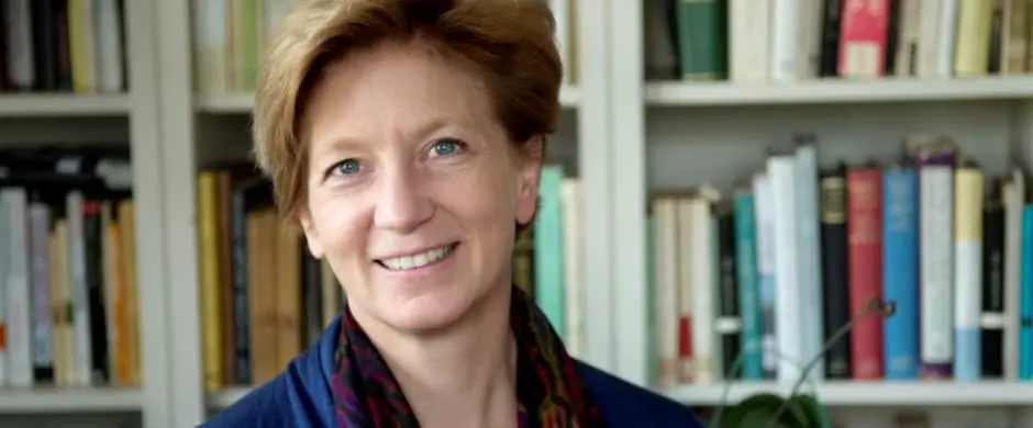 A smiling woman with short light-brown hair stands in front of bookshelves filled with various books. She is wearing a dark top and a patterned scarf. The setting appears to be an office or library.