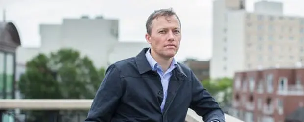 A man wearing a dark jacket and blue shirt stands outdoors on a balcony, with blurred buildings and trees in the background on a cloudy day.