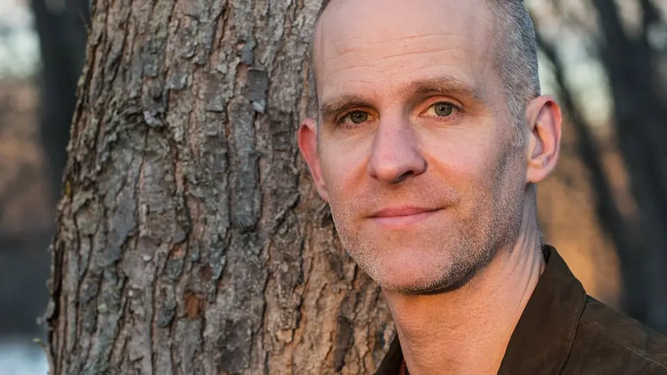 A middle-aged man with short, light hair and a slight smile stands close to a tree trunk outdoors. The background is softly focused, showing more trees and natural light.