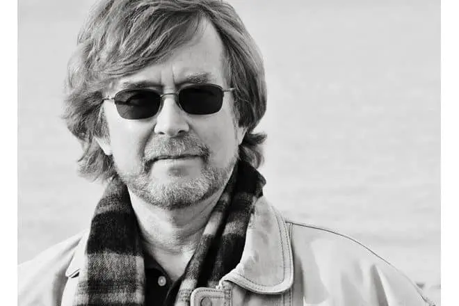 A man with medium-length hair and a beard, wearing dark sunglasses, a plaid scarf, and a light jacket, stands outdoors. The background is blurred and appears to be a bright, open area.