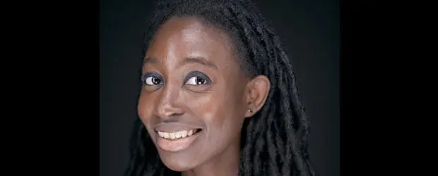 A woman with long, dark, twisted hair smiles at the camera. She has dark skin, wears subtle makeup, and is set against a plain black background.