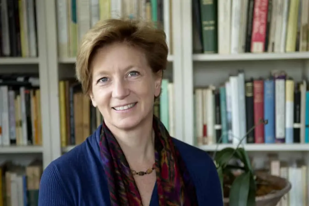 A woman with short light brown hair, wearing a blue top and patterned scarf, smiles in front of a bookshelf filled with books and a potted plant.