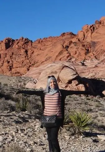 A woman wearing a hijab and striped shirt stands in a desert landscape with her arms outstretched, red rock formations and clear blue sky in the background.