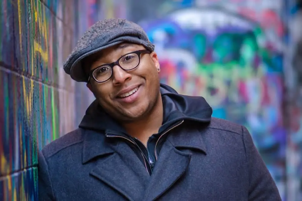 A smiling person wearing glasses, a gray flat cap, and a dark coat stands against a graffiti-covered wall in an alleyway.
