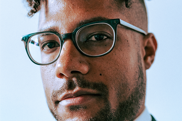 A close-up of a man wearing black-rimmed glasses, with short curly hair, facial hair, and a serious expression, looking directly at the camera against a light background.