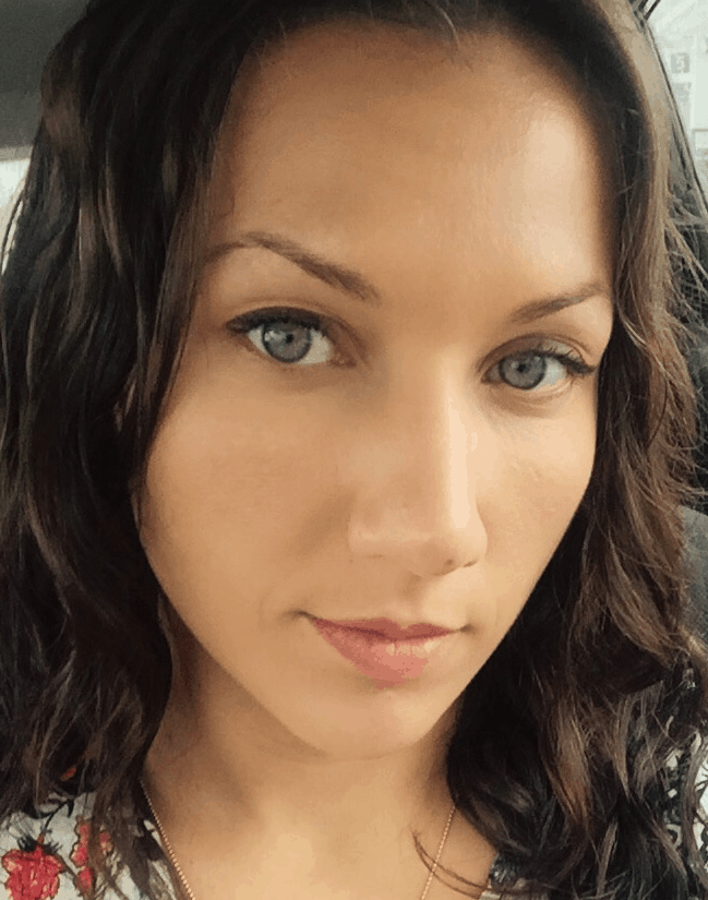 A woman with wavy brown hair and blue eyes looks directly at the camera with a neutral expression, sitting inside a car.