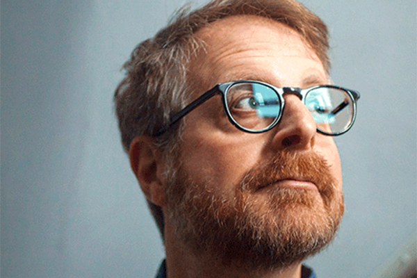 A man with light brown hair, a beard, and glasses looks slightly upward and to the side, against a light blue background.