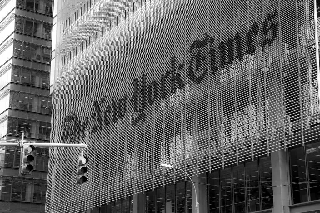 The New York Times building facade in black and white, with the newspaper’s name prominently displayed in large letters on the exterior mesh. A traffic light hangs in the foreground.