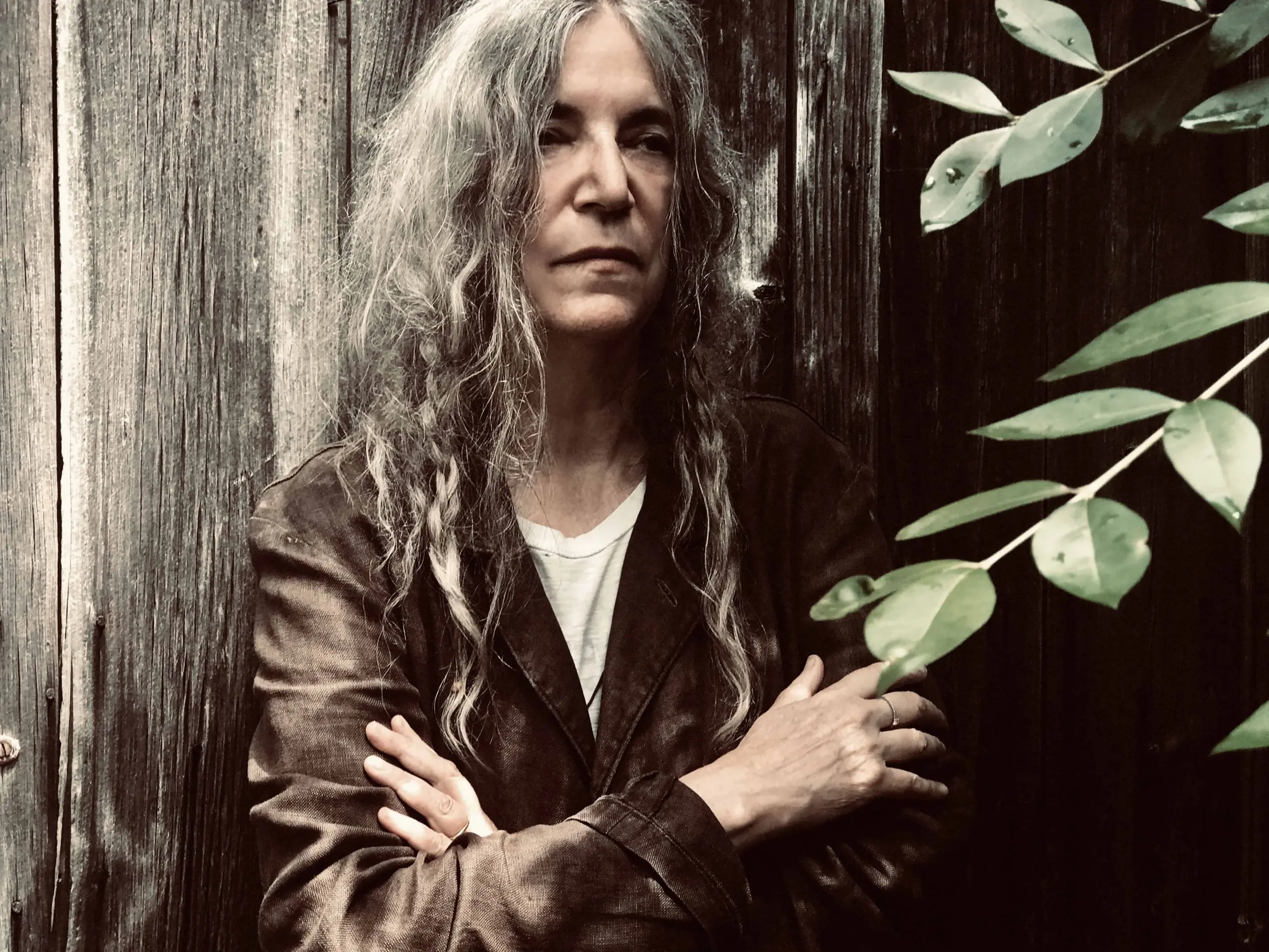 A person with long, wavy gray hair stands with arms crossed in front of a weathered wooden wall, wearing a brown jacket and white shirt—reminiscent of portraits from the 2020 PEN America Literary Gala. Green leaves partially frame the right side.