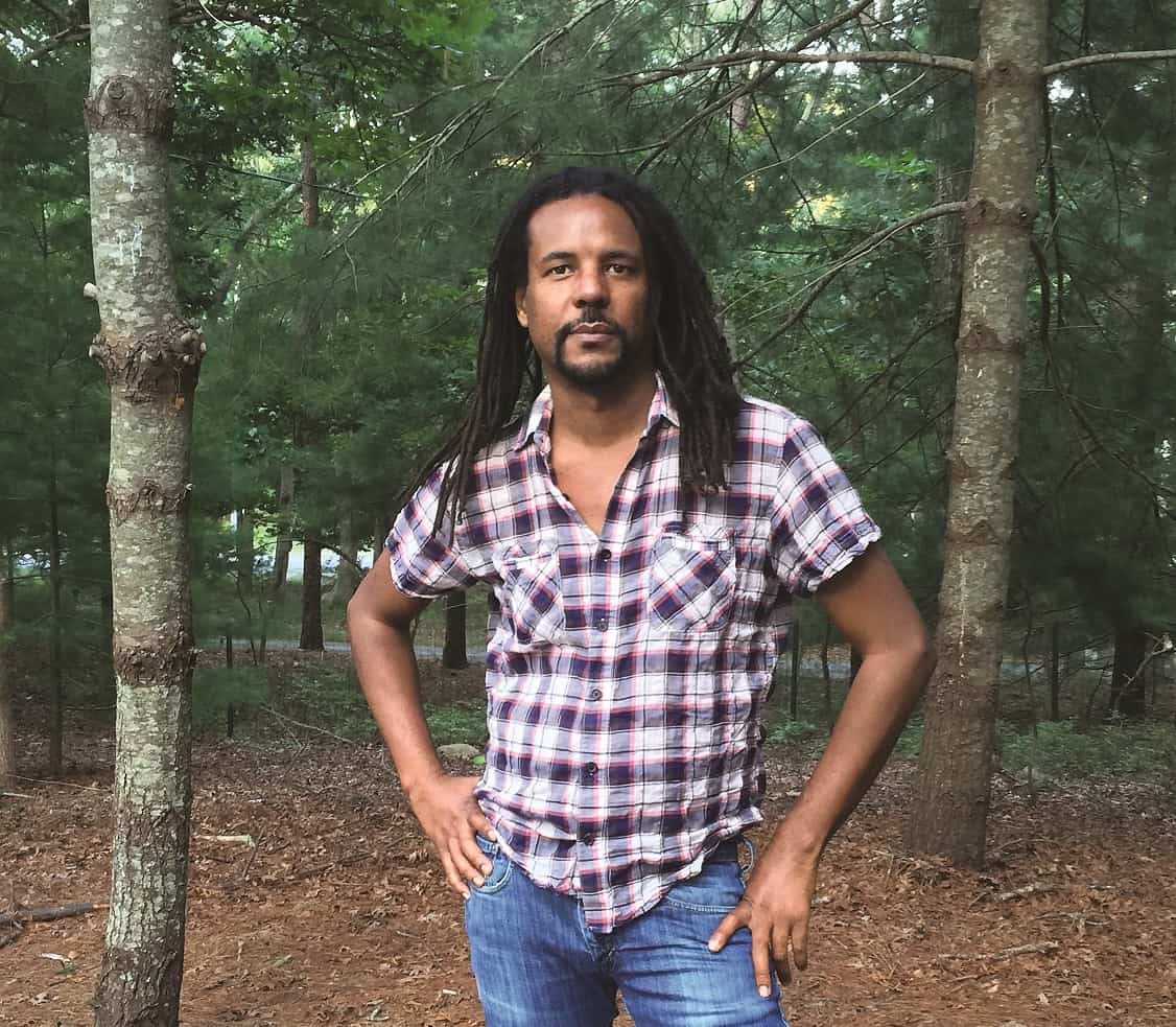 A man with long dreadlocks in a plaid shirt and jeans stands among tall trees in a wooded area, looking at the camera with one hand on his hip.