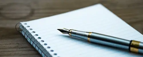 A silver fountain pen rests on an open spiral notebook with lined paper, placed on a wooden surface.