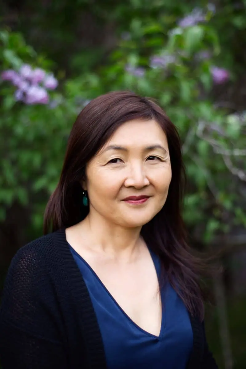 A woman with long dark hair and a blue top stands outdoors in front of blurred greenery and purple flowers, looking at the camera with a gentle smile.
