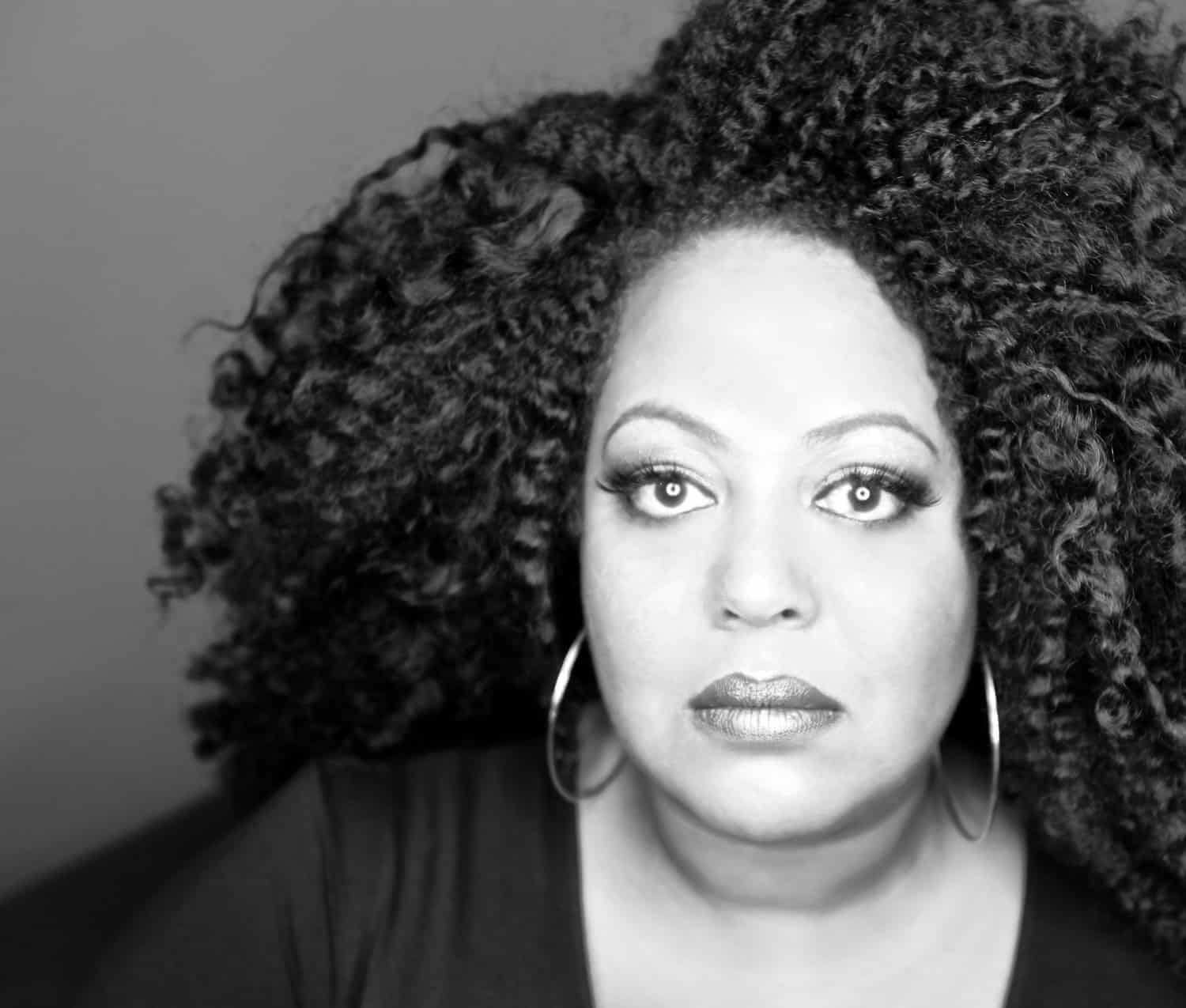 Black and white portrait of a woman with voluminous curly hair, large hoop earrings, and a serious expression, looking directly at the camera.