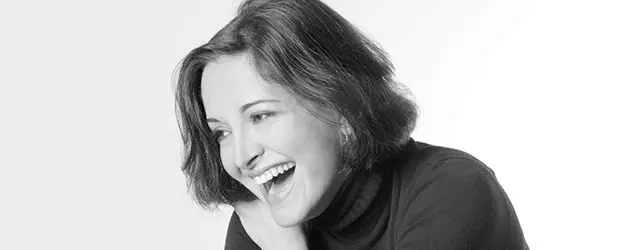 A woman with short hair wearing a dark turtleneck smiles and laughs, looking to the side. The photo is in black and white with a light background.