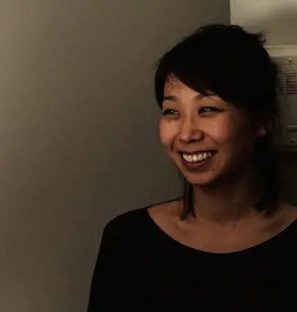 A woman with dark hair smiles and looks to the side, standing against a plain wall in dim lighting.