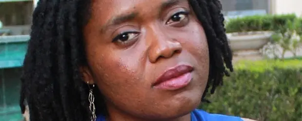 A woman with dark skin and black dreadlocks wearing a blue top and dangling earrings looks at the camera with a neutral expression. There is greenery and a turquoise structure in the blurred background.