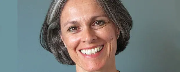 A woman with short, gray and dark hair smiles warmly at the camera. She is wearing small pearl earrings and a dark top, with a plain blue background behind her.