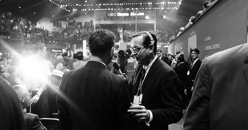 A black-and-white photo shows a busy convention floor with a well-dressed reporter interviewing a man in a suit. Large crowds fill the seats, and bright stage lights shine as an ABC News sign hangs in the background.