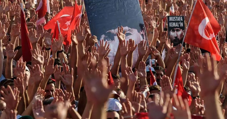 A large crowd of people with raised hands holds Turkish flags and posters, including one with Mustafa Kemal Atatürk’s face. Many wear red and white, creating a sea of color and unity.