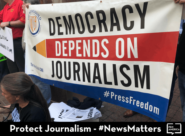 People hold a banner reading “Democracy Depends on Journalism #PressFreedom” at a rally. Text at the bottom says, “Protect Journalism – #NewsMatters” with the PEN America logo in the corner.