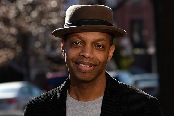 A man wearing a brown fedora and a dark jacket smiles at the camera outdoors, with blurry trees and cars in the background.