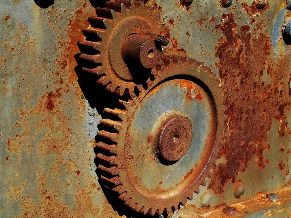Two rusty metal gears interlock against a corroded, weathered metal surface, showing signs of age and oxidation with various shades of brown and orange rust patches.