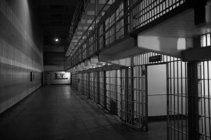 A black and white photo of a prison corridor with metal bars and cells on the right, and an empty hallway stretching into the distance on the left. The area is dimly lit, creating a stark and somber atmosphere.