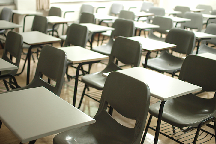 Rows of empty gray chairs with attached white desks are arranged neatly in a classroom with sunlight coming through the windows in the background.