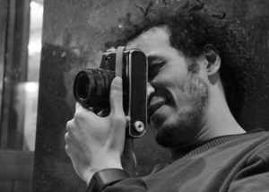 A person with curly hair holds a vintage film camera up to their eye, appearing to take a photograph. The image is in black and white, and the person is smiling slightly.